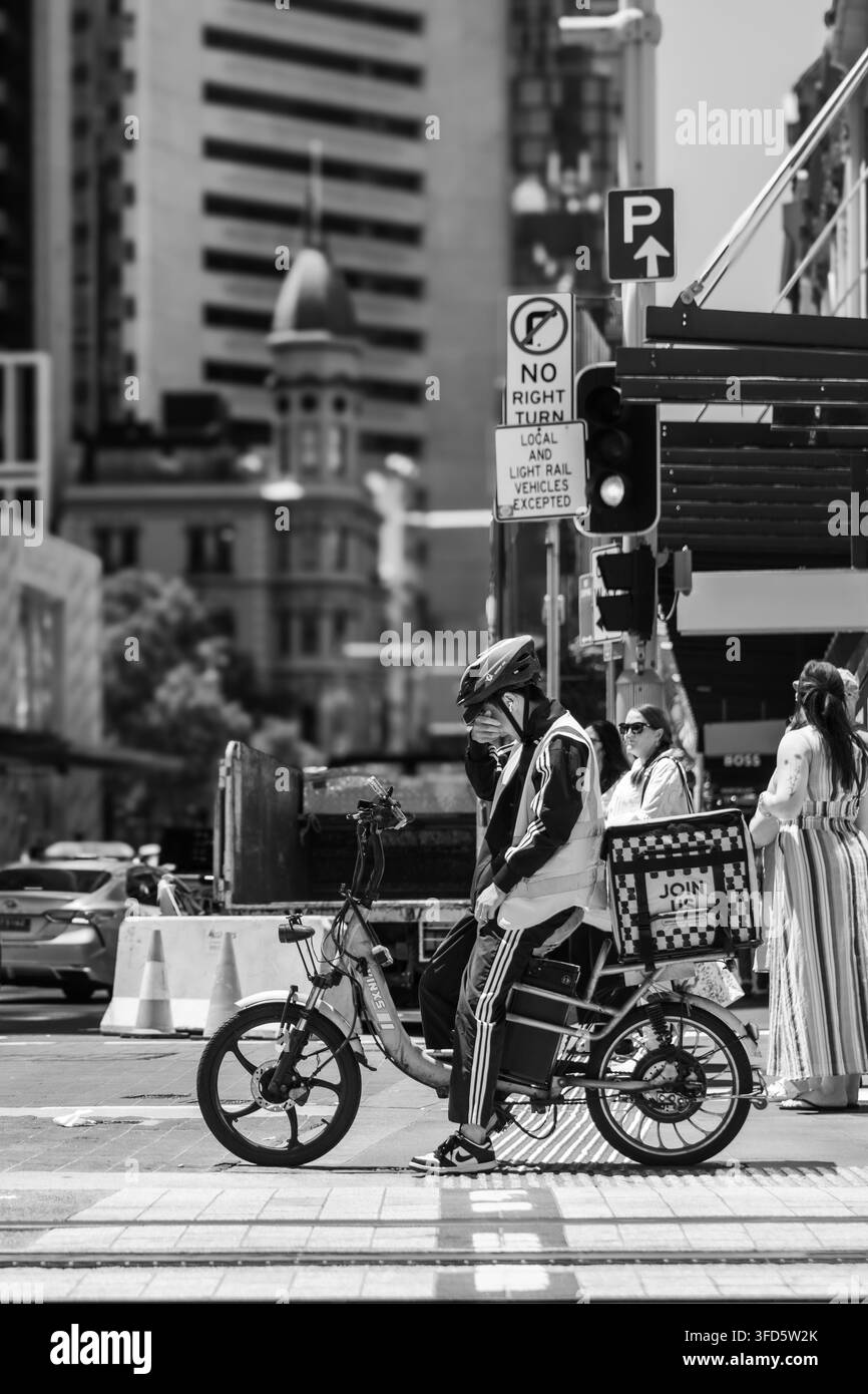 Ein Lieferfahrer auf einem E-Bike wartet an einer Stadtkreuzung, umgeben von Verkehrsschildern, Fußgängern und hohen Gebäuden. Stockfoto