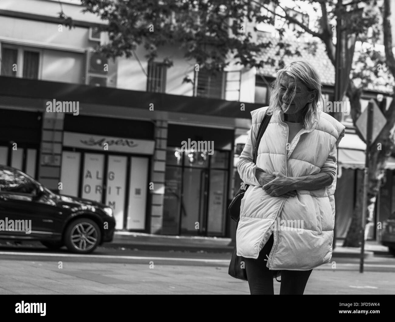 Ältere, obdachlose Frau in einer Daunenweste, die an Geschäften und einem geparkten Auto in der Stadt vorbeigeht, AlltagsSzene im städtischen Leben. Stockfoto