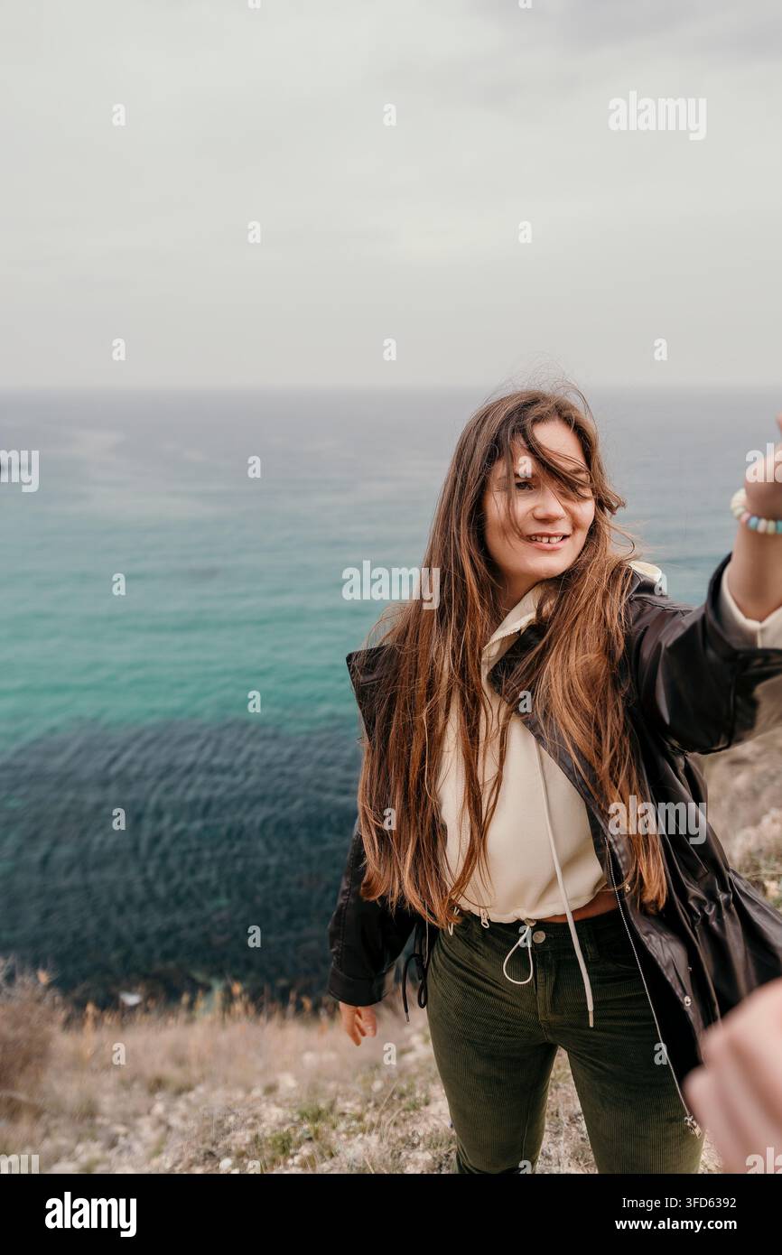 Frau Klippen Ozean lächelnde Frau mit langen Haaren steht auf einer Klippe mit Blick auf ein türkisfarbenes Meer an einem windigen Tag Stockfoto