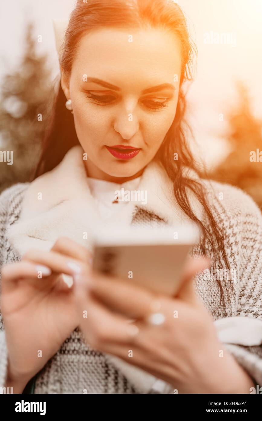 Frau im Freien, die ein Smartphone verwendet, während sie einen stilvollen Mantel trägt, während der goldenen Stunde mit warmem Sonnenlicht Stockfoto