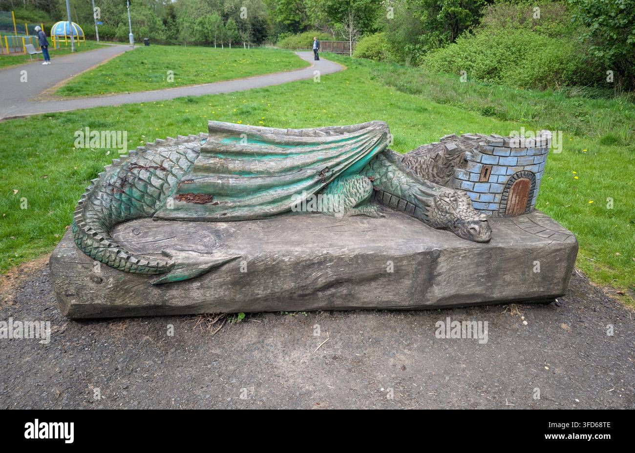 Eine detaillierte Holzskulptur eines schlafenden grünen Drachen, der sich um einen Miniatur-Schlossturm schlingt, dient als skurrile Bank in einem öffentlichen Park. Stockfoto