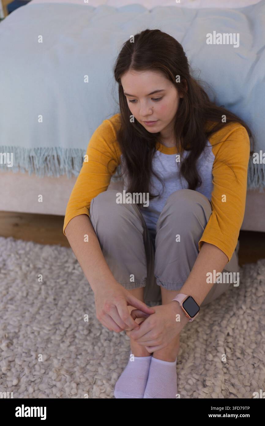 Teenagerin sitzt auf texturierter Wolldecke neben dem Bett, passt ihre Zehen an, trägt ein Senfhemd und eine Smartwatch Stockfoto