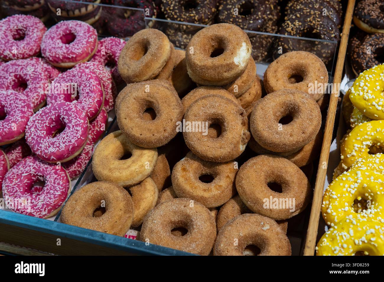 Reihen von Zimtzucker-, rosa glasierten und gelb glasierten Donuts sind in einer Bäckereivitrine zusammengestapelt. Stockfoto