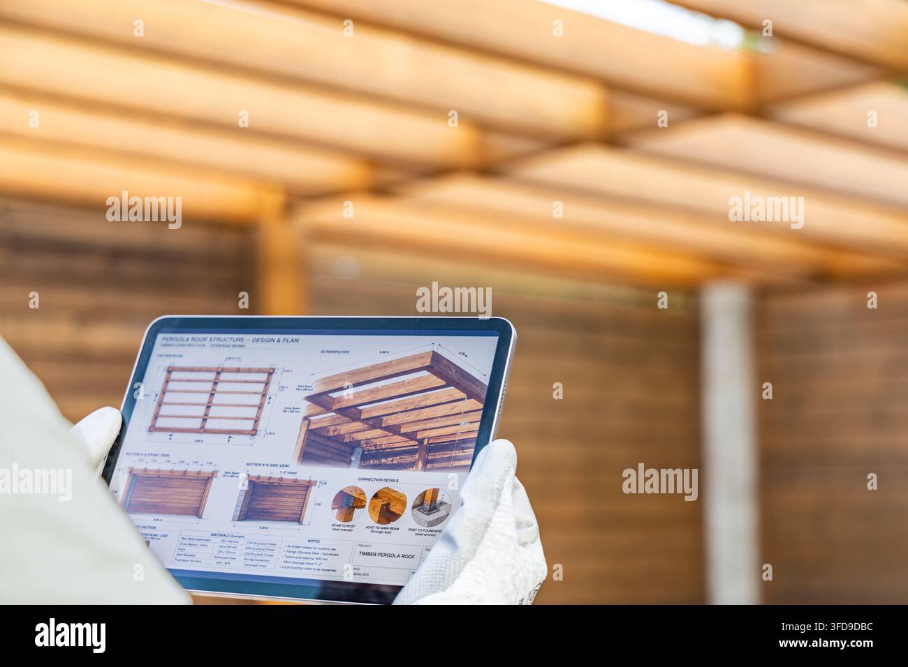 Eine Person mit Handschuhen hält ein Tablet, das detaillierte Baupläne und 3D-Visualisierungen einer hölzernen Pergola-Struktur anzeigt Stockfoto