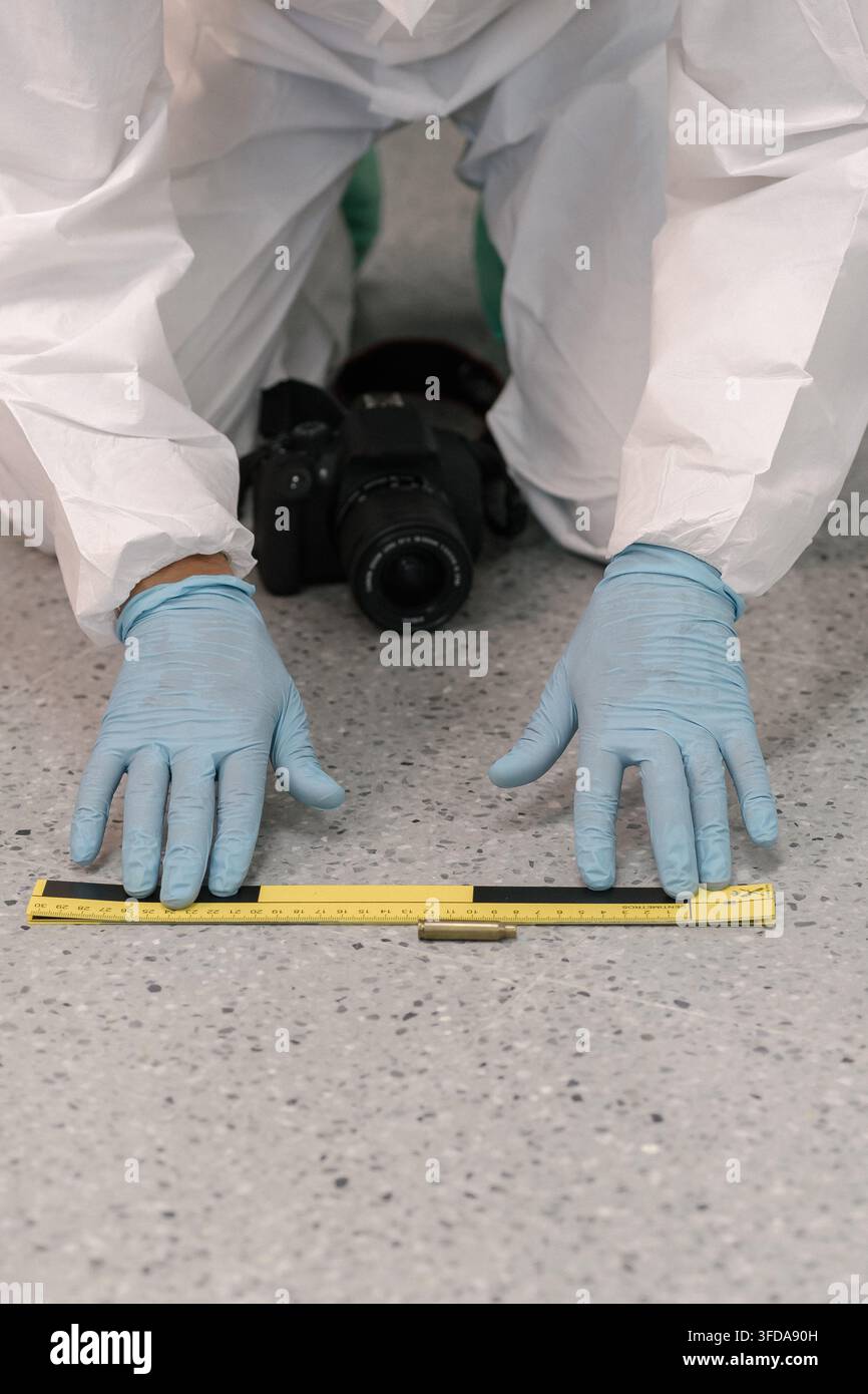 Ein Forensiker positioniert eine gelbe Messskala neben einem kleinen Objekt auf dem Terrazzoboden, wobei eine Kamera zwischen den Knien ruht. Stockfoto