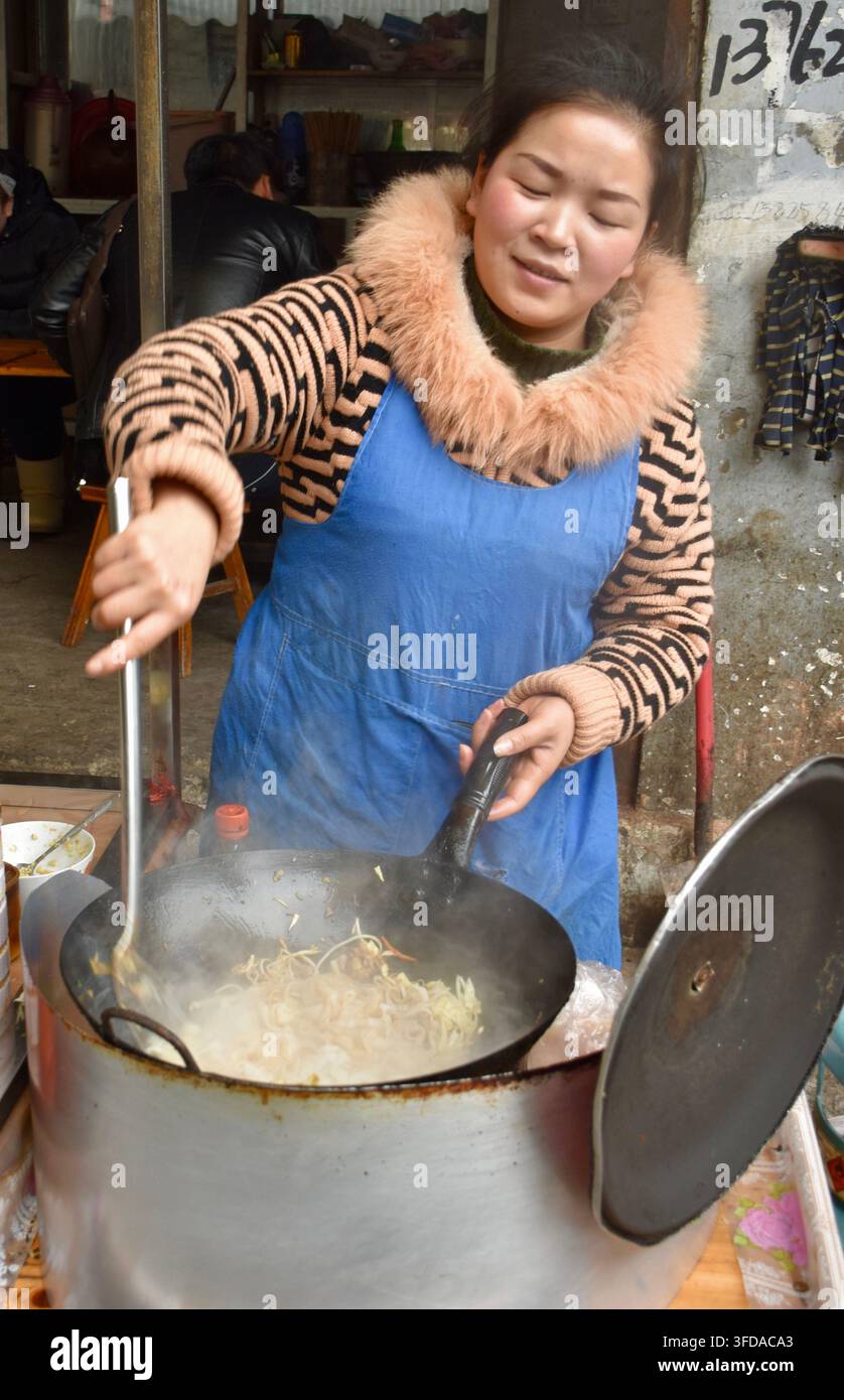 Guizhou, China - 14. März 2017: Verkäuferin kocht authentisches Straßenfood in einem großen schwarzen Wok, trägt eine blaue Schürze und einen Wintermantel mit Kunstpelzkragen Stockfoto