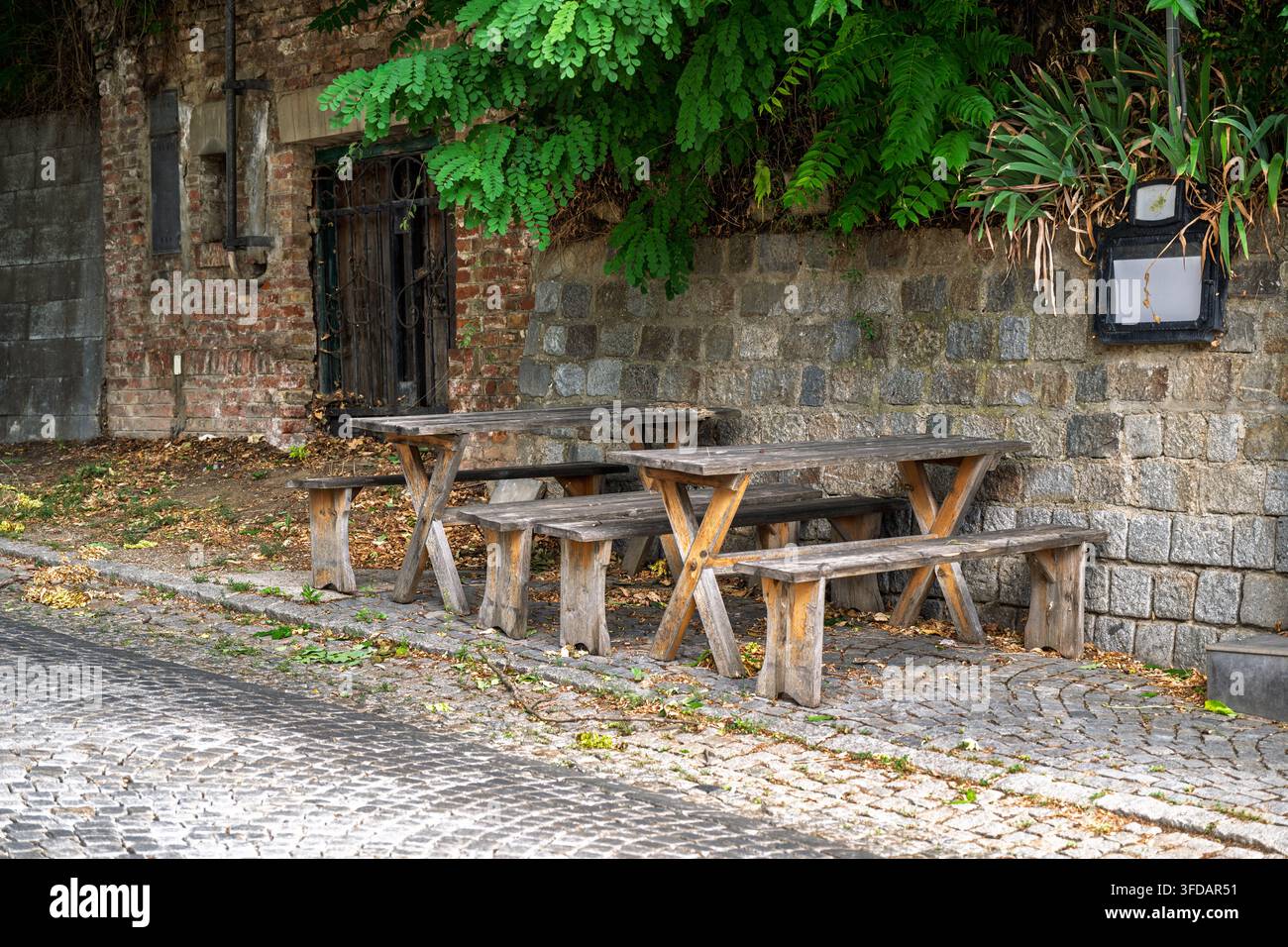 Zwei rustikale Holztische stehen leer auf einem stillen Kopfsteinpflasterweg und laden Besucher ein, eine Pause einzulegen und die charmante, schattige Atmosphäre entlang dieses Weges zu genießen Stockfoto