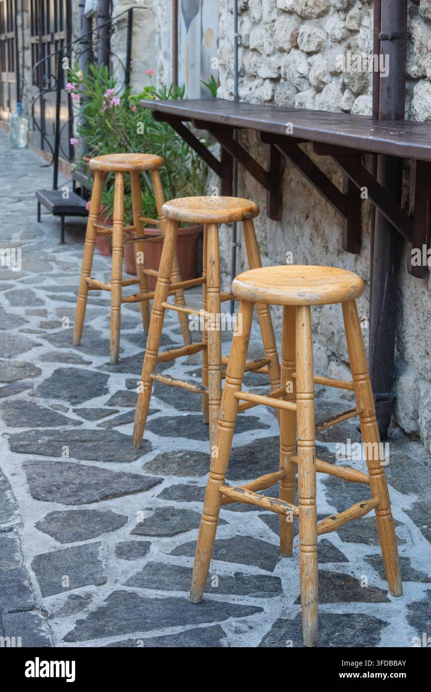 Drei hohe Holzbarhocker stehen in einer Reihe unter einem an der Wand montierten Außentresen entlang einer rustikalen steinplätzigen Gasse im Tageslicht. Stockfoto