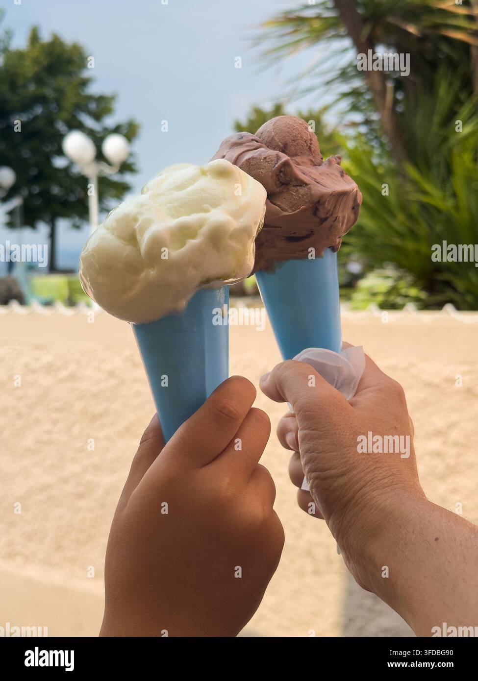 Hände halten Vanille- und Schokoladeneisbecher in einer Gartenumgebung mit Washingtonia filifera. Die Szene steht für Urlaub, Familie und Genuss Stockfoto