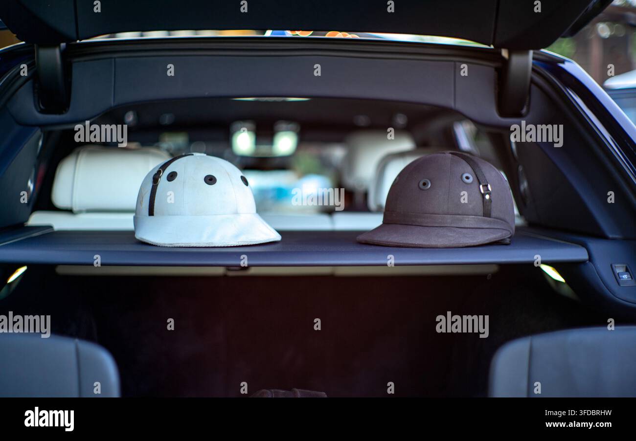 Ein klares horizontales Foto mit einem weißen und einem schwarzen Reitsport-Polohelm nebeneinander auf dem Fach im offenen Kofferraum eines Luxuswagens Stockfoto