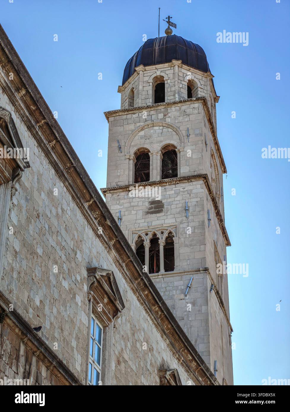 Eine atemberaubende niedrigwinklige Aufnahme fängt den alten steinernen Glockenturm und die architektonischen Details des Franziskanerklosters unter klarem Himmel ein. - Smartphone-aufgenommenes Stockfoto