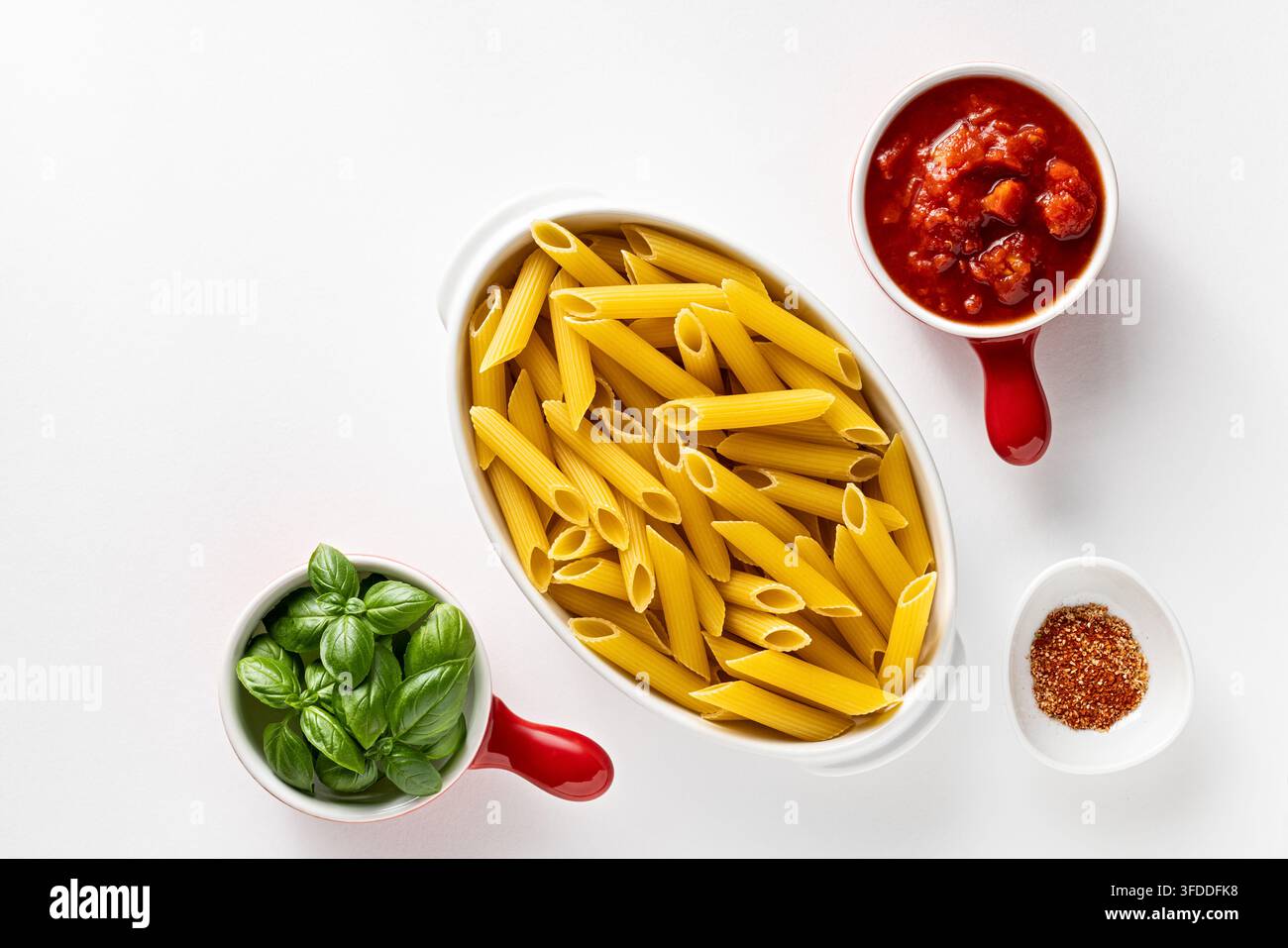 Diagonale Flat Lay von trockener Penne-Pasta mit Basilikum-Tomatensauce und Gewürzen in einer sauberen minimalen Food-Anordnung Stockfoto
