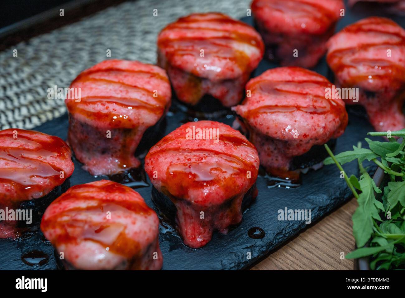 Eine Ansicht von oben zeigt mehrere gebackene Sushi-Rollen, die mit einer rosa cremigen Sauce und süßer Sozanglasur belegt sind, die auf einem dunklen Schieferteller neben frischem Grünes angeordnet sind Stockfoto