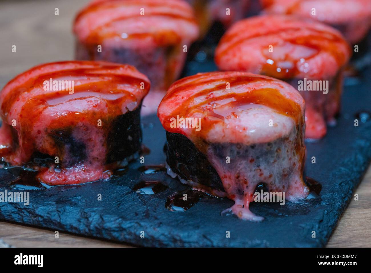 Eine Nahaufnahme zeigt mehrere gebackene oder angegrillte Sushi-Rollen, die mit einer rosa cremigen Sauce und süßer Sozuglasur belegt sind und auf einem dunklen Schieferteller arrangiert wurden. Stockfoto