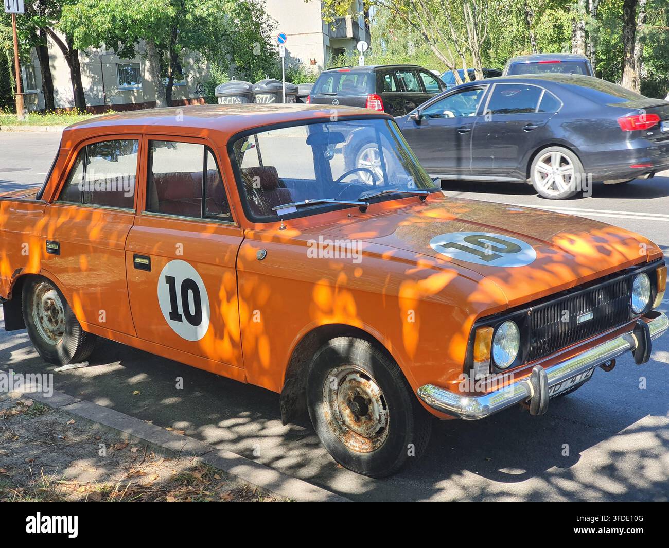 Eine vintage orangefarbene Limousine aus der Sowjetzeit mit weißen runden Rennnummer "10"-Aufklebern auf der Fahrertür und der Motorhaube, geparkt in einer sonnigen Stadt Stockfoto
