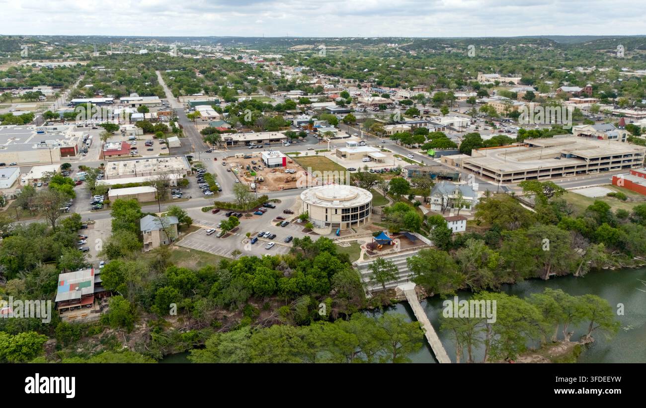 Luftbilddrohnenaufnahme über die Innenstadt von Kerrville mit Gewerbegebäuden, Straßen, einem Flussuferpark, einer Fußgängerbrücke und bewaldeter Landschaft. Stockfoto