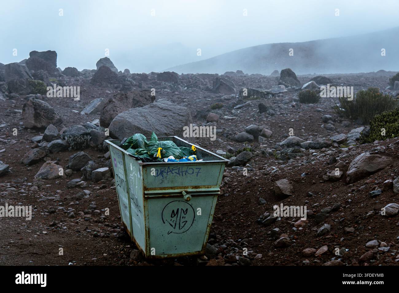 Grüner Müllcontainer, der mit Abfall gefüllt ist und auf einem felsigen, nebligen Berghang steht, betont die Umweltverschmutzung Stockfoto