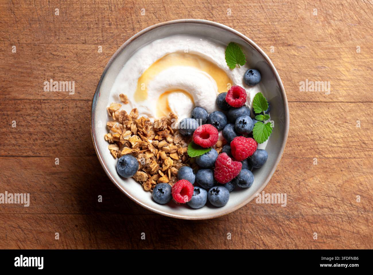 Frühstücksschüssel mit griechischem Joghurt, belegt mit Granola, gemischten Beeren und einem Tropfen Honig. Granola-Schüssel mit Blaubeeren und Himbeeren auf Holzunterlage Stockfoto