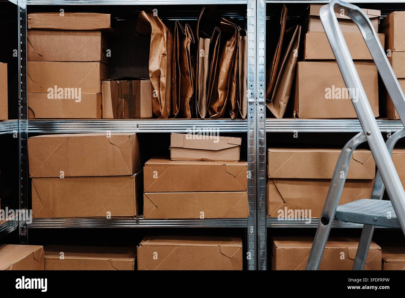Kartons und Kraftpapiertüten füllen Metallregale in einem Lagerraum neben einer Trittleiter. Recycelbare Verpackungen unterstützen Nachhaltigkeit Stockfoto