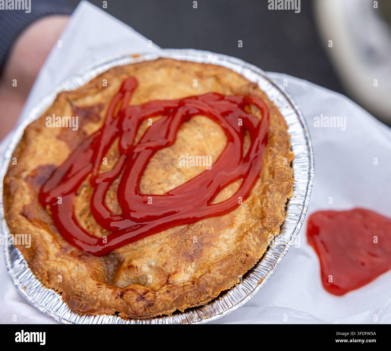 Die ausgewählte Region zeigt eine traditionelle, goldbraune Einzel-Fleischpastete, die in einer Folienform serviert wird und mit einer großzügigen Spirale Tomatenketchup gekrönt ist. Stockfoto