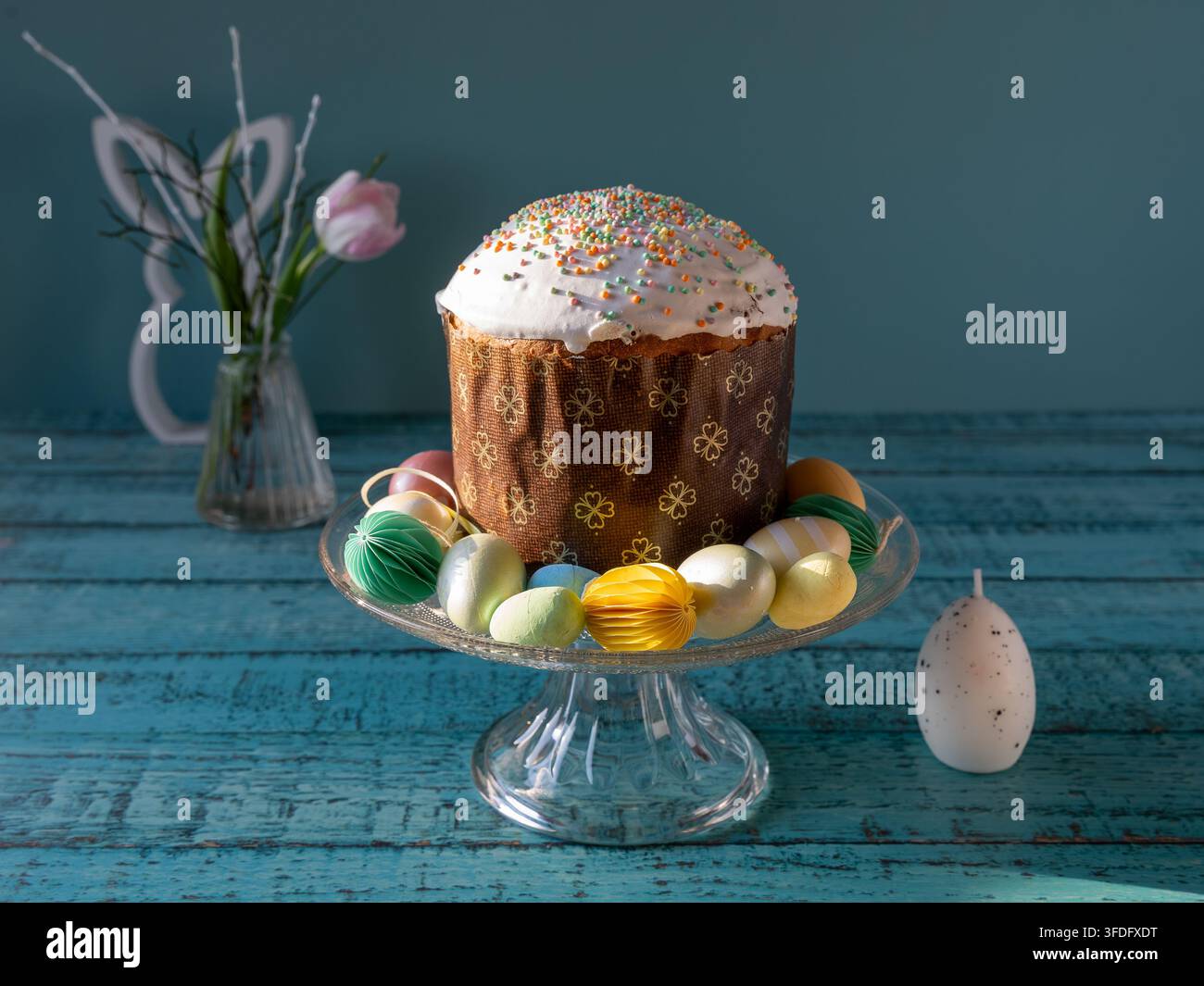 Traditionelle osteuropäische Paska, auch bekannt als Pasca, auf einem gläsernen Kuchenständer präsentiert, umgeben von bunten dekorativen Eiern, Tulpen und Frühlingsdekoration Stockfoto