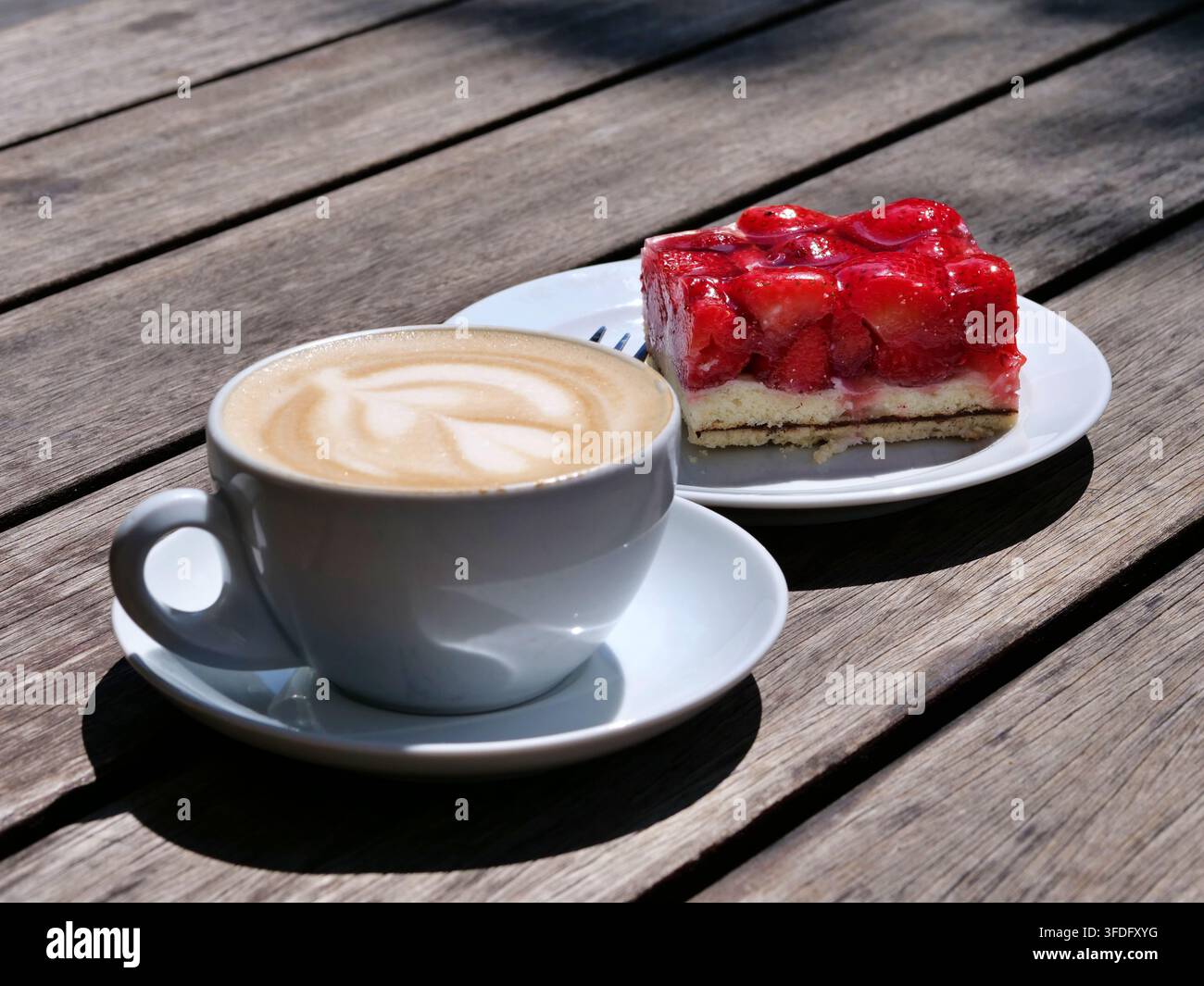 Hochwinkelaufnahme zeigt eine weiße Tasse mit warmem Cappuccino mit detaillierter Latte Art neben einem Stück frischem Erdbeerkuchen mit roter Glasur. Serviert auf Stockfoto