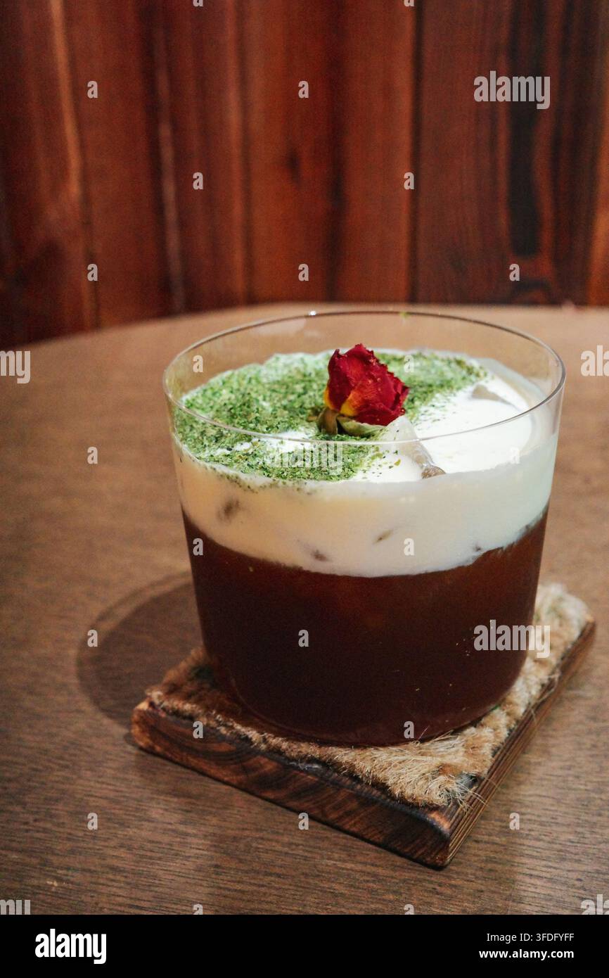 Der dunkelbraune Latte im durchsichtigen Glas ist mit weißem Milchschaum bedeckt. Der Milchschaum ist mit hellem Matcha-Pulver bestreut, und ein dunkelroter Dr. Stockfoto