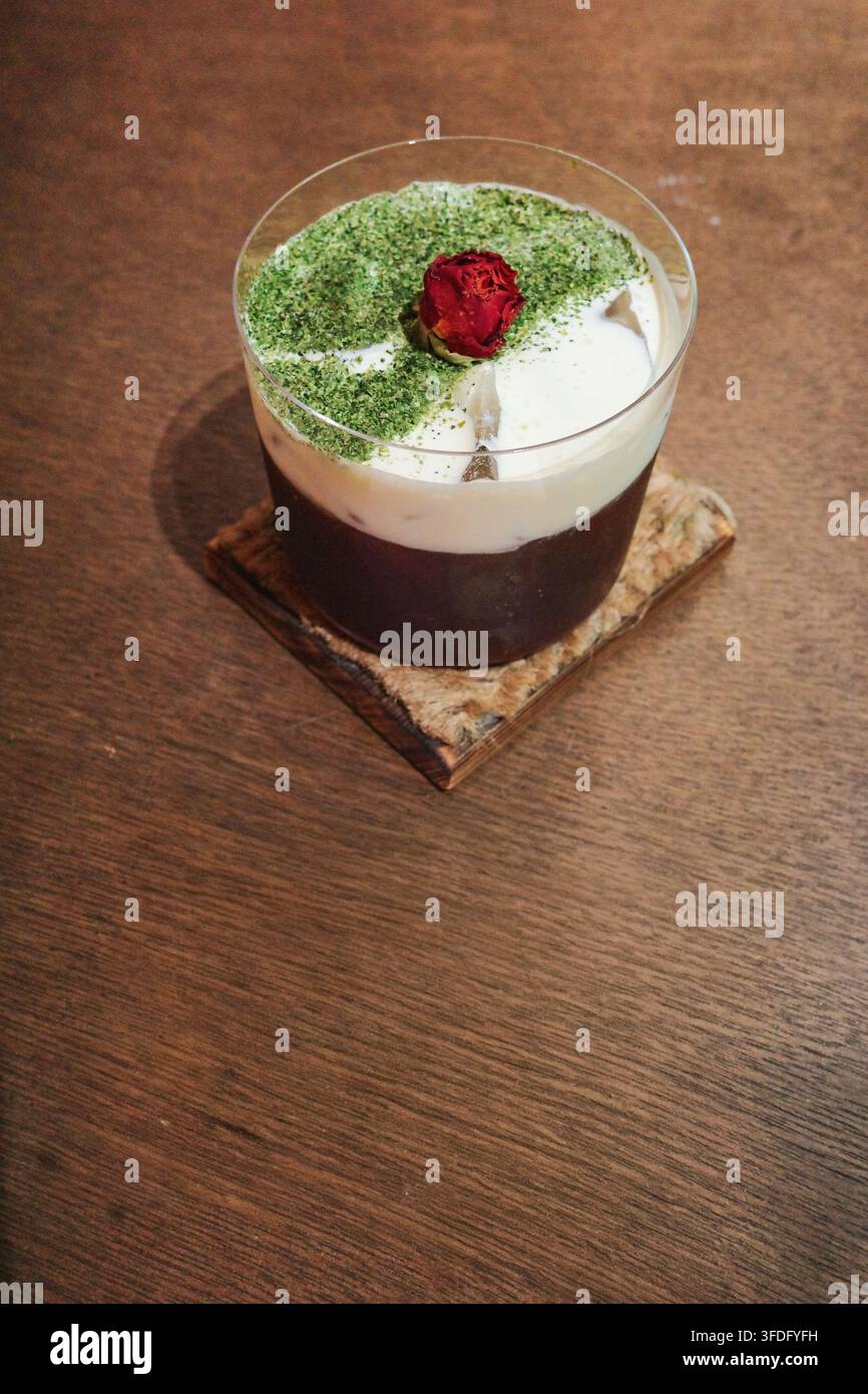 Eine transparente Glas Tasse auf einem quadratischen Leinenuntersetzer enthält dunklen Kaffee und Milchschaum. Die Oberseite ist mit hellem grünem Matcha-Pulver bestreut, und der ce Stockfoto
