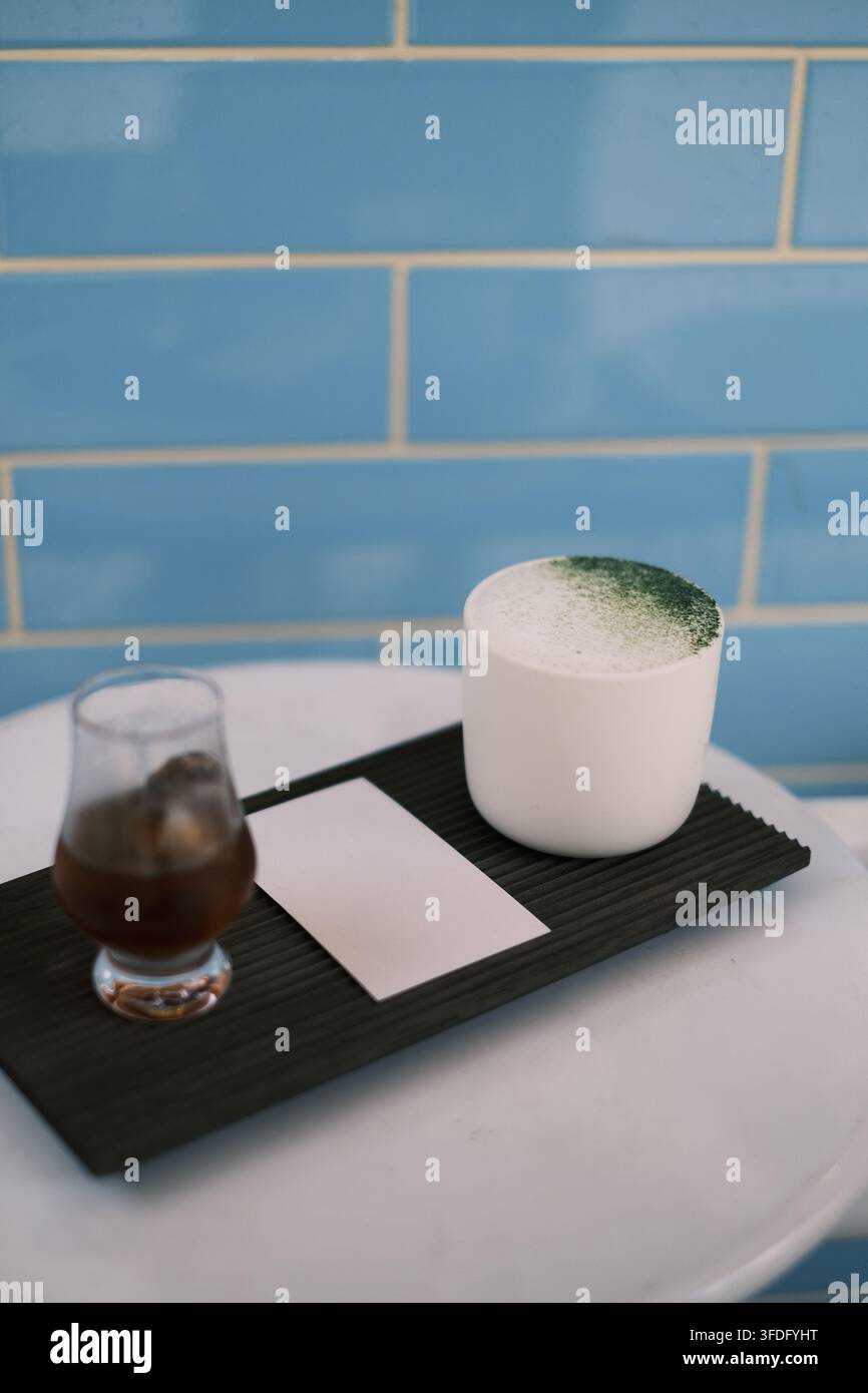 Eine kurze weiße Porzellantasse enthält einen milchig weißen Matcha-Latte, mit smaragdgrünem Matcha-Pulver oben drauf gestreut. Das kleine Glas daneben enthält ein i Stockfoto