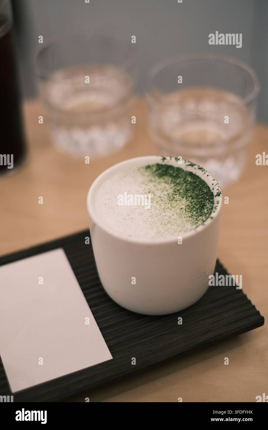 Eine kurze weiße Porzellantasse enthält einen milchig weißen Matcha-Latte, auf dessen Oberfläche dunkelgrünes Matcha-Pulver gestreut ist. Die Tasse ruht auf schwarzem Holz Stockfoto