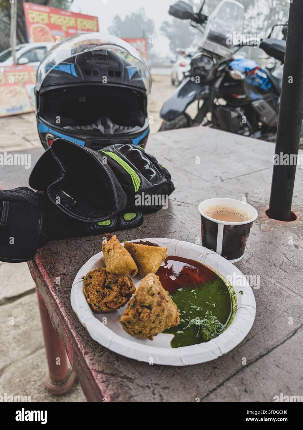 Jan1st2026 Dehradun Indien. Motorradhelm und Handschuhe neben einem Teller mit indischen Snacks und Tee auf einem Tisch am Straßenrand mit Motorrädern im Hintergrund Stockfoto