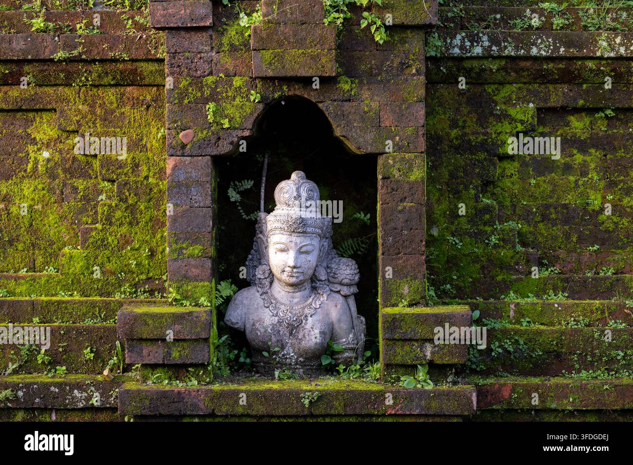 UBUD, BALI, INDONESIEN 10. MÄRZ 2026 Ein Götterkopf ist von einer dunklen Nische und moosbewachsenem traditionellem Ziegelmauerwerk eingerahmt. Stockfoto