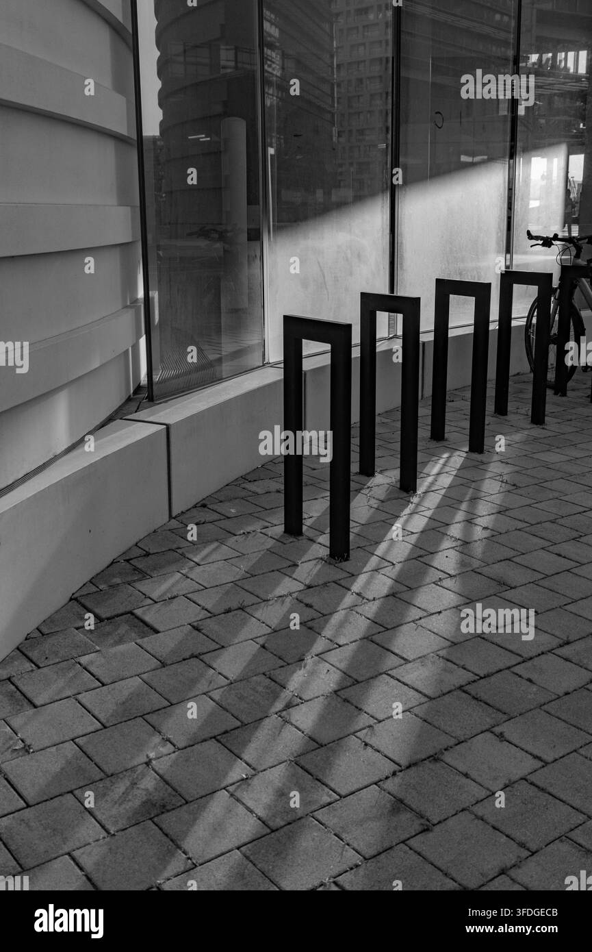 Schwarz-Weiß-Bild auf Straßenniveau von leeren geometrischen Fahrradständern vor einem Glasbürogebäude, mit langen Schatten und Stadtreflexionen Stockfoto