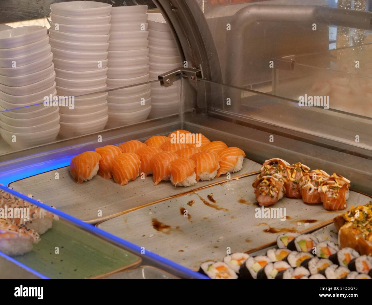 Schrägaufnahme eines gekühlten Restaurant-Vitrinens, das frischen Lachs-Nigiri, Maki-Rollen und Gewürzschalen zeigt, beleuchtet von subtilem blauem LED-Licht Stockfoto
