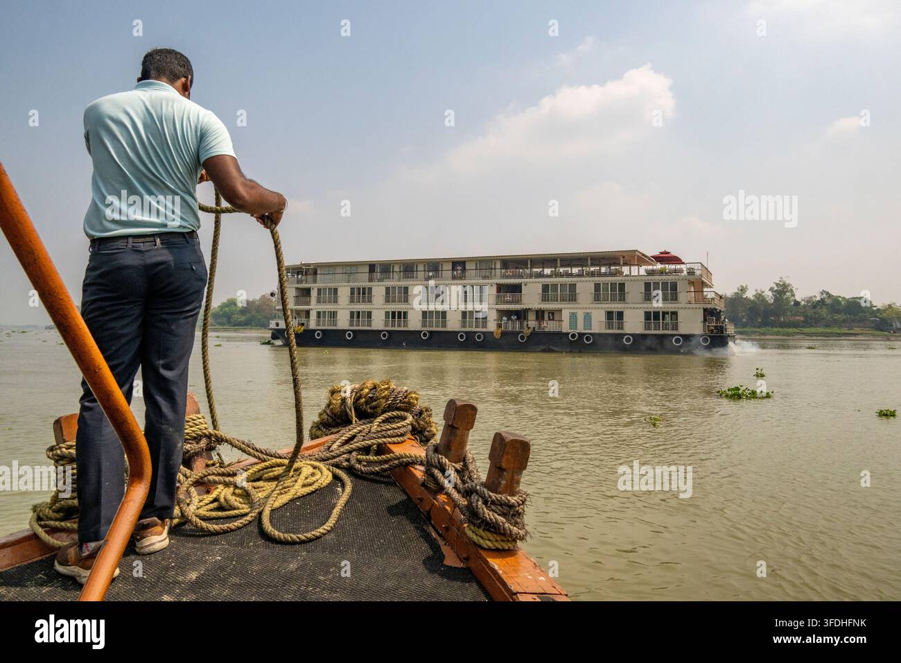 Ein Mann sichert ein Seil am Bug eines kleinen Bootes, Sampan oder Landboot, während ein großes Flusskreuzfahrtschiff auf den trüben Gewässern des Ganges vorbeifährt Stockfoto