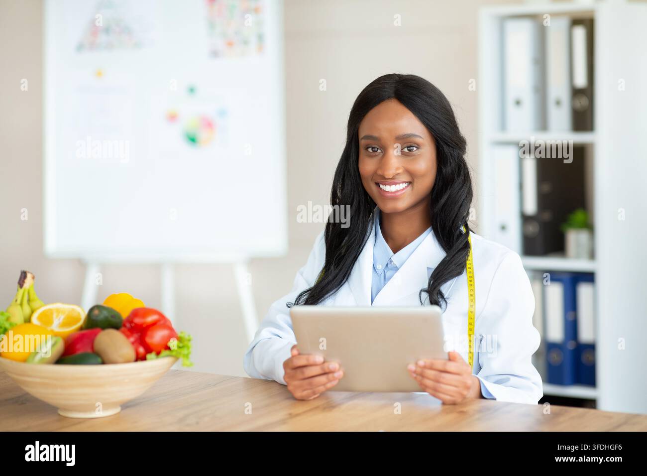 Junger schwarzer Ernährungsberater mit Touchpad, der einen pflanzlichen Speiseplan für den Kunden in der Klinik erstellt, Leerraum. Fröhliche weibliche Diätetikerin mit Tablettenkonsulti Stockfoto