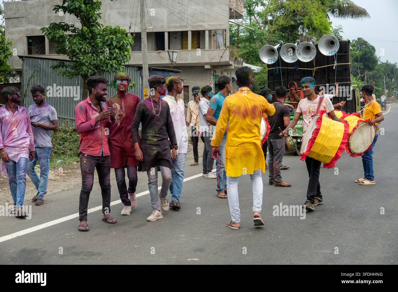 Junge Männer, die mit farbigem Pulver bedeckt sind, tanzen und spielen Trommeln während einer lebhaften Straßenfeier des hinduistischen Festivals Holi Stockfoto