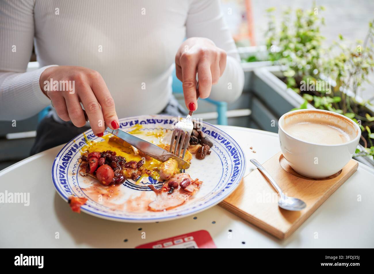 Nahaufnahme einer Frau, die ein herzhaftes Frühstück in einem Café isst, Wurst mit Eiern, Bohnen und Pilzen schneidet, Cappuccino in der Nähe, Konzept des Alltagslebens, Ernährung, Essgewohnheiten Stockfoto