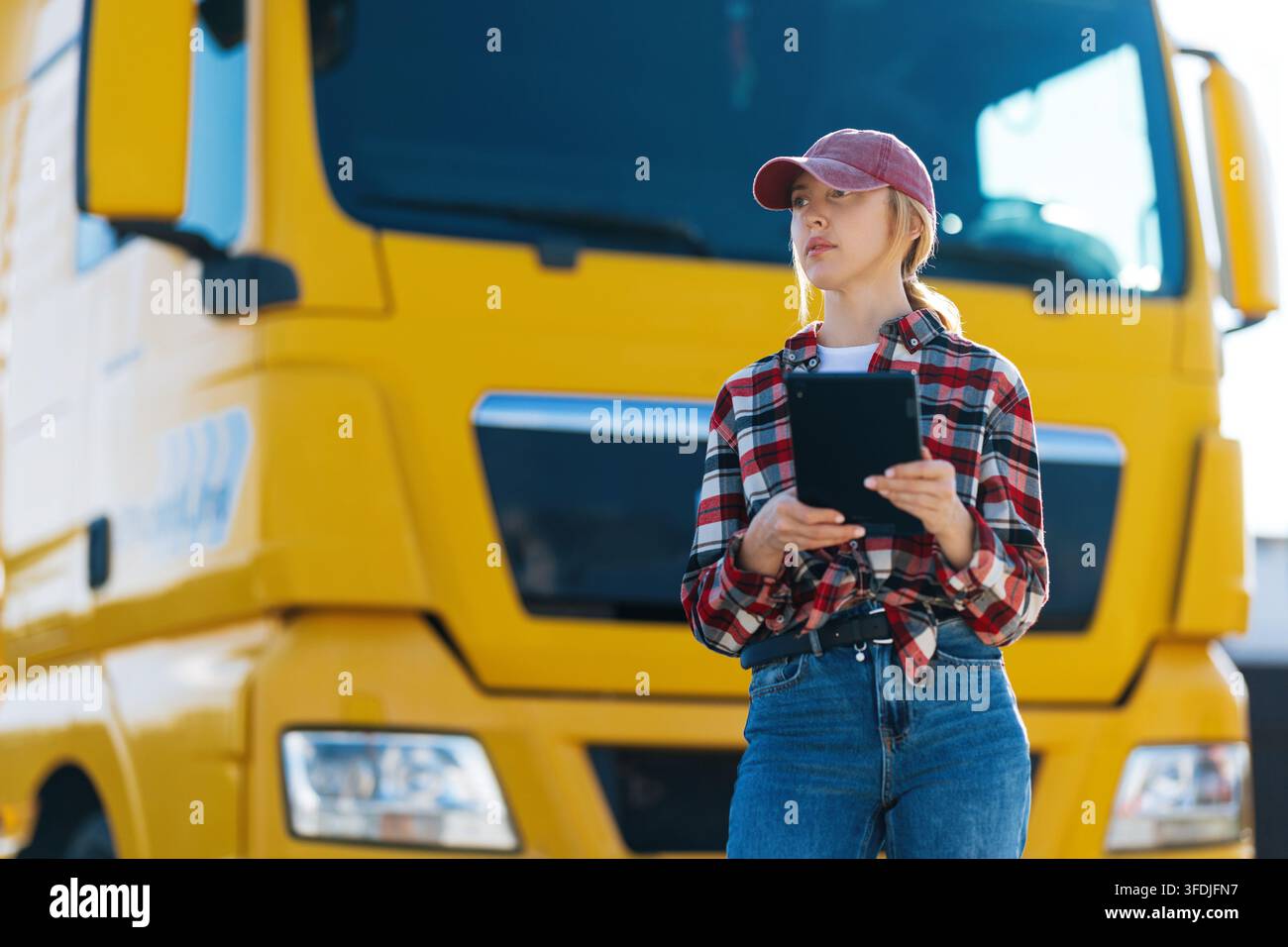 Eine Frau mit Baseballkappe und kariertem Hemd hält ein Tablet, während sie neben einem großen gelben Sattelschlepper steht Stockfoto