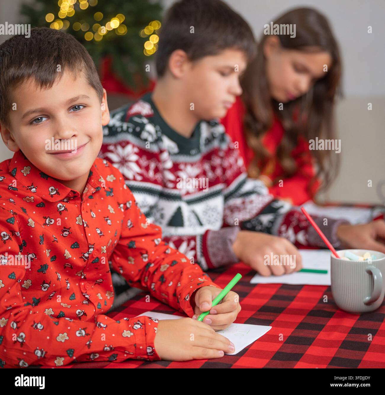 Drei Kinder in Weihnachtspyjamas zeichnen am Tisch mit festlicher Beleuchtung, mit Kindern, Junge, Lächeln, Geschwisterdetail Stockfoto