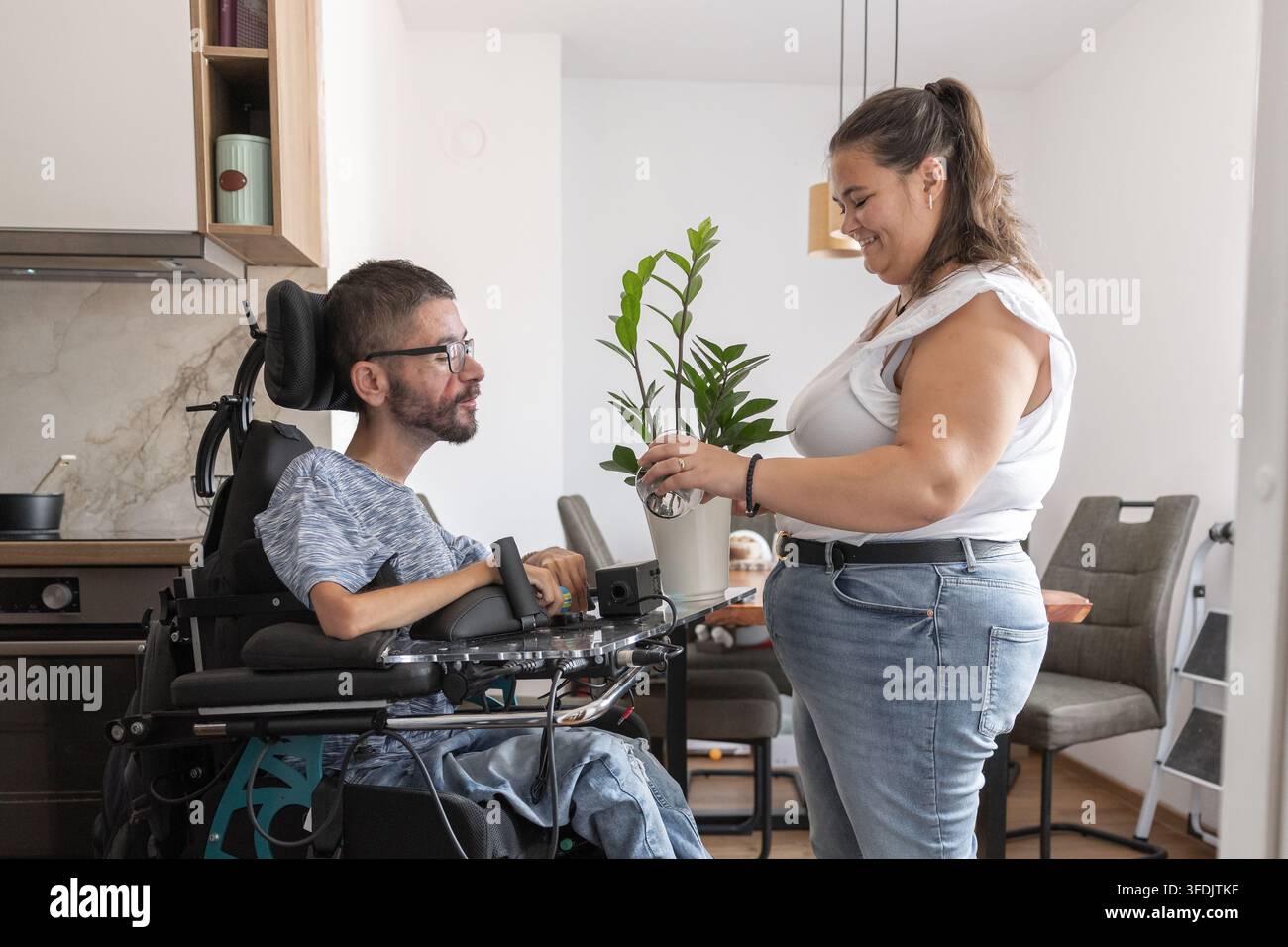 Persönliche Assistentin und ein Mann mit Behinderung pflegen gemeinsam eine Zimmerpflanze und genießen ihren Duft zu Hause. Stockfoto