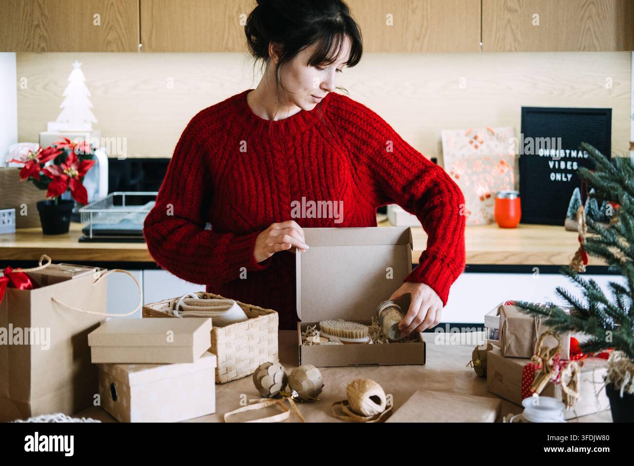 Frau legt Glas in offene Kraftbox mit Pinsel in der Küche in der Nähe des Schilds „CHRISTMAS VIBES ONLY“. Überprüfung des Weihnachtsgeschenksets vor dem Versand, Spa-Geschenkbox Stockfoto