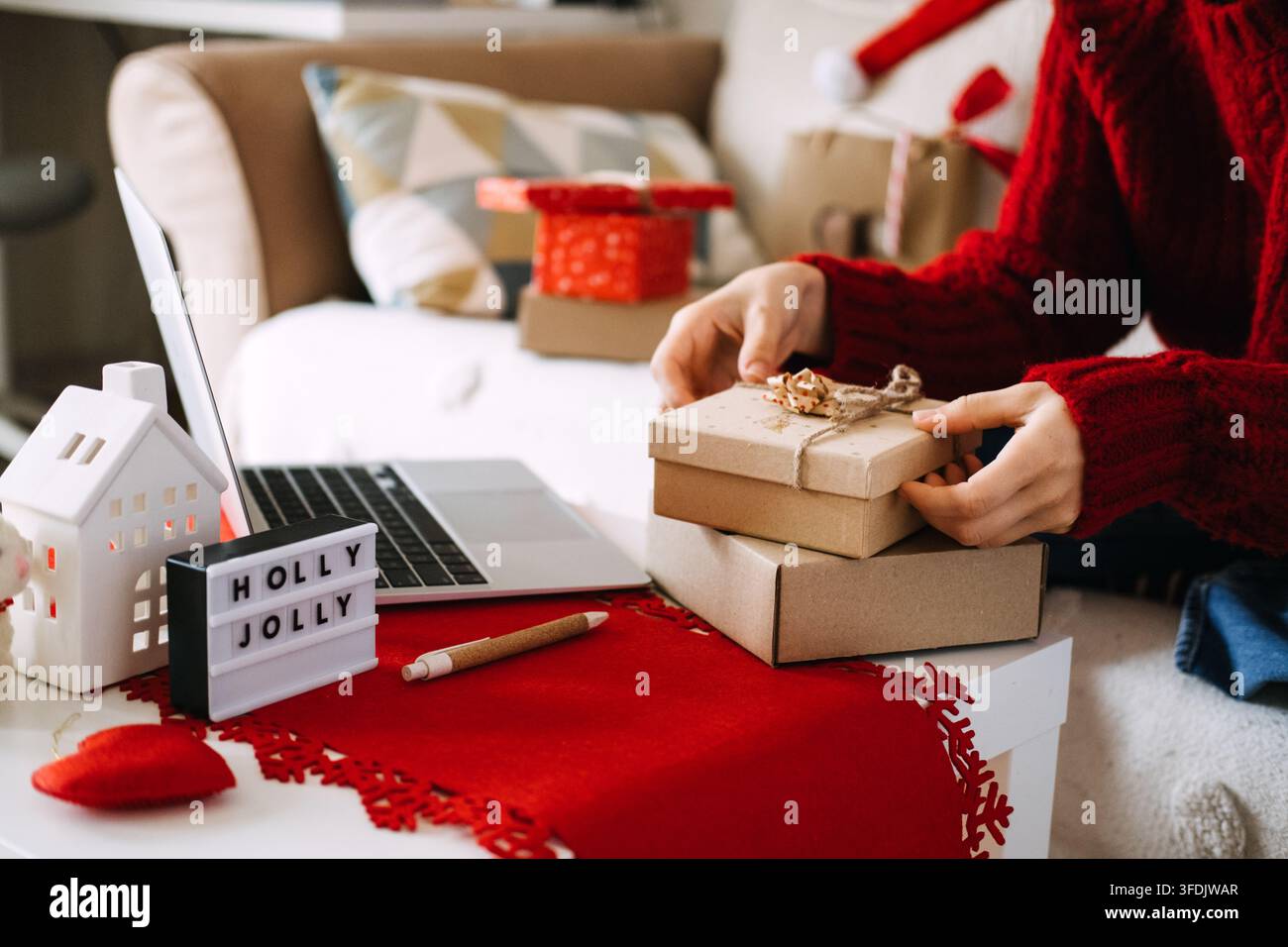 Hände stapeln Kraftkisten neben einem Laptop auf dem Tisch mit einem HOLLY JOLLY-Schild und einem Keramikhaus. Rückgabe eines unerwünschten Weihnachtsgeschenks, Geschenkaustauschrichtlinie, Post Stockfoto