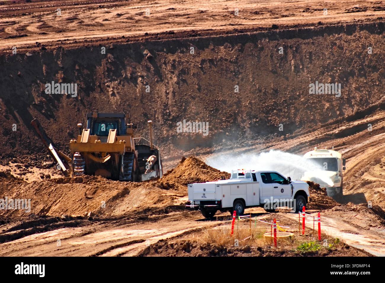 Ein Wassertankwagen sprüht Staubbindendes Sprühwasser über eine aktive Ausgrabungsstelle, während ein Bulldozer und ein Nutzfahrzeug zwischen freigelegtem Boden und Verkehrskoni arbeiten. Stockfoto