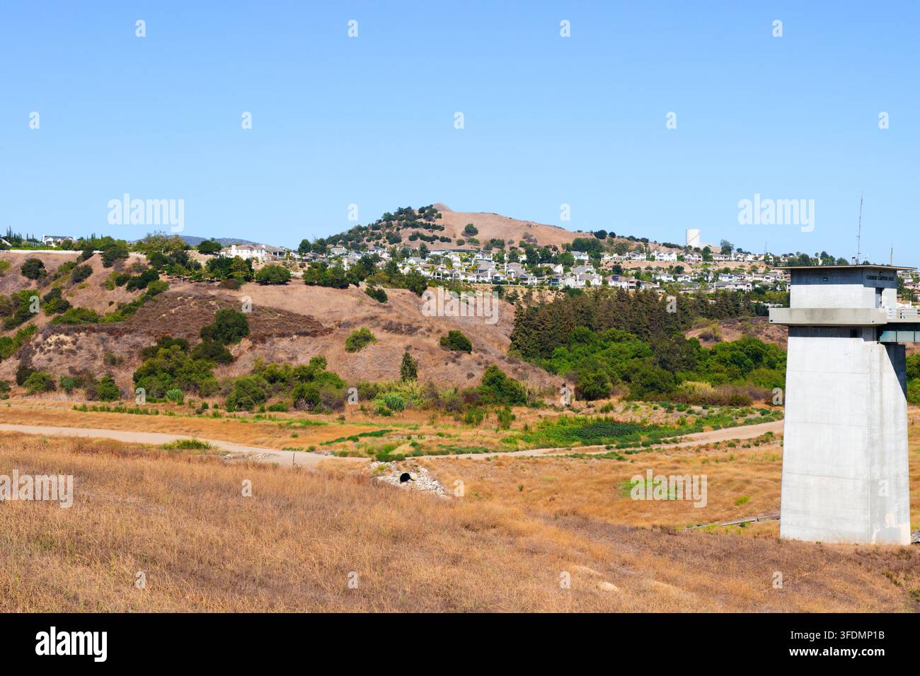 Ein Betondamm-Kontrollturm erhebt sich über ein trockenes Überflutungsbecken, mit chaparral-bewachsenen Hügeln und Vorstadthäusern unter einem klaren blauen Sommerhimmel. Stockfoto