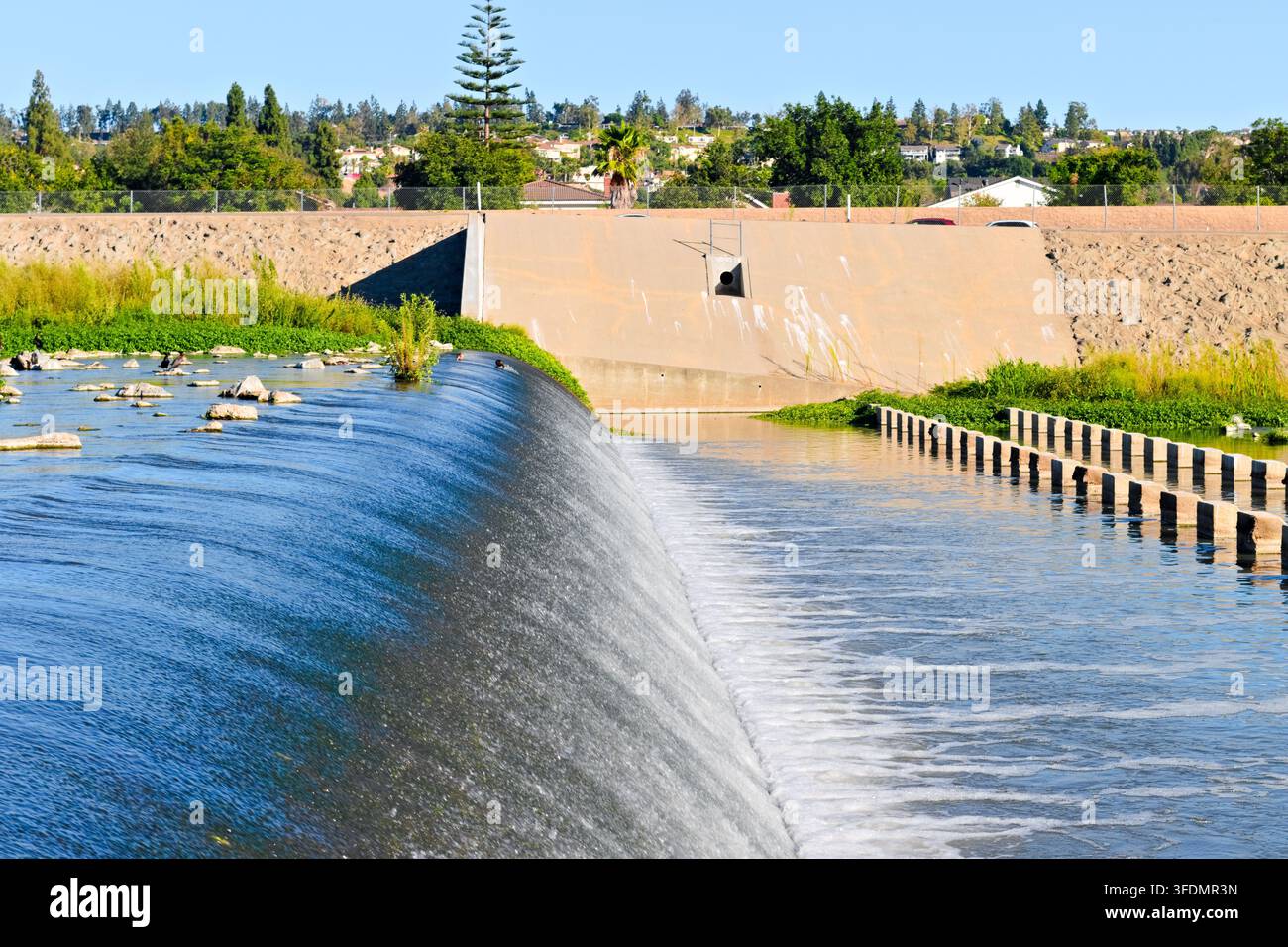 Wasser fließt über einen gebogenen Betonüberlauf in einen breiten Kanal, der von Hochwasserschutzbauwerken und grüner Vegetation umgeben ist. Stockfoto
