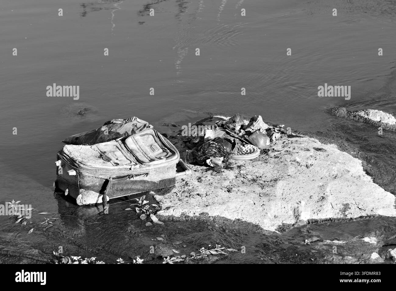 Verlassene Taschen, Kleidung und anderer Abfall liegen auf einer Betonplatte neben ruhigem Flusswasser und veranschaulichen Müll und Verschmutzung in einem städtischen Gewässer. Stockfoto