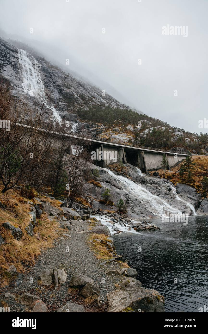 Der majestätische Langfossen-Wasserfall in Norwegen stürzt einen steilen, dunklen felsigen Berghang hinunter unter einem nebligen, bewölkten Himmel, mit einer modernen Autobahnbrücke c Stockfoto