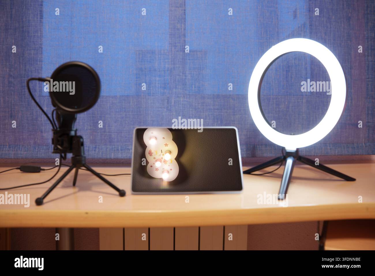 Podcast-Mikrofon und Ringlicht neben einem Tablet, das ein festliches Schneemann-Bild zeigt, auf einem Holzschreibtisch mit blauem Vorhang als Hintergrund, wodurch ein gemütliches Studio entsteht Stockfoto