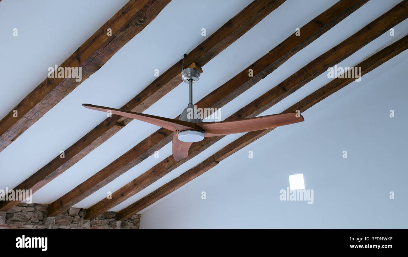 Eine nach oben gerichtete, schräge Ansicht zeigt einen modernen Holzdeckenventilator vor einer weißen gewölbten Decke, die von markanten dunklen Holzbalken durchzogen ist, hochl Stockfoto